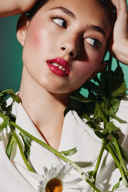 Close-up of a young woman with glossy red lipstick and dewy makeup wearing a white shirt, holding green leaves and a white flower. Concept of beauty, fashion, and creative portrait.