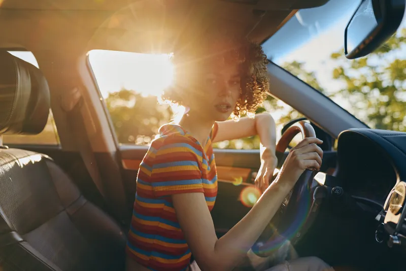 Young woman with curly hair drives a car at golden hour, wearing a colorful striped shirt, and smiles softly as sunlight streams through the windshield, creating a relaxed road trip mood.