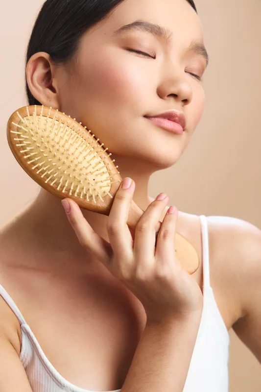 asian woman enjoys a relaxing grooming moment, brushing her hair with a wooden brush, eyes closed, serene smile, soft lighting, calm atmosphere, beauty routine in a peaceful personal care setting.