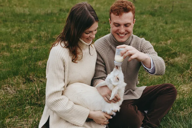 Young couple in cozy sweaters enjoys feeding a baby goat with a bottle while sitting on grass in a relaxed outdoor lifestyle moment.