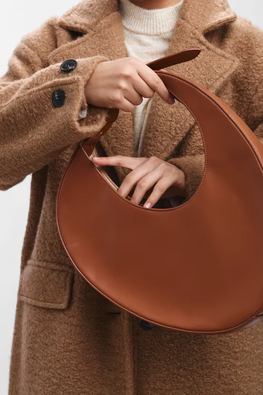 asian woman wearing a beige camel coat holds a circular leather bag, showcasing fashion elegance and contemporary style in a minimalist setting.