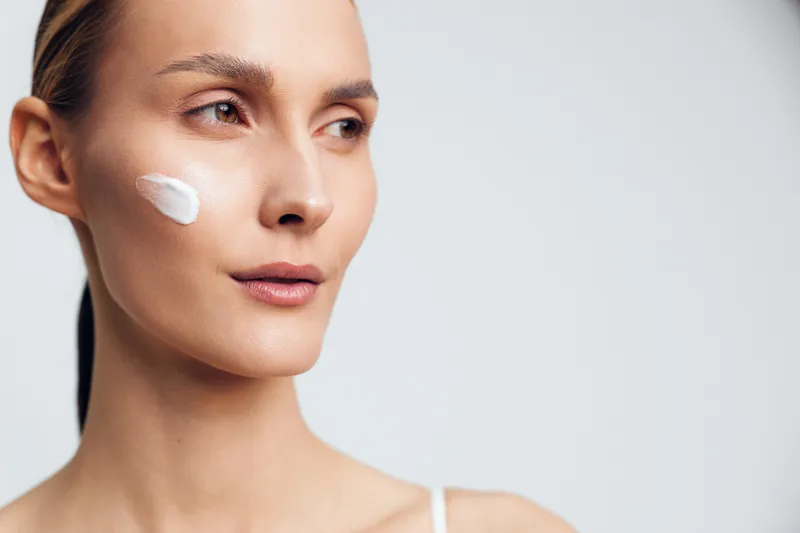 Woman applies white face cream on cheek in a close up beauty portrait, skincare and self care concept, calm expression, soft studio lighting on light background