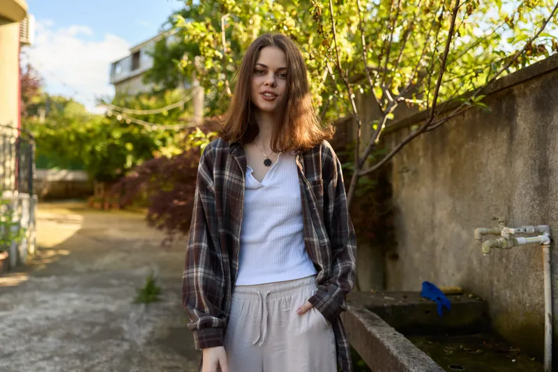 Smiling woman in plaid overshirt and white tee stands outdoors among greenery, presenting a casual lifestyle portrait with natural daylight and relaxed mood