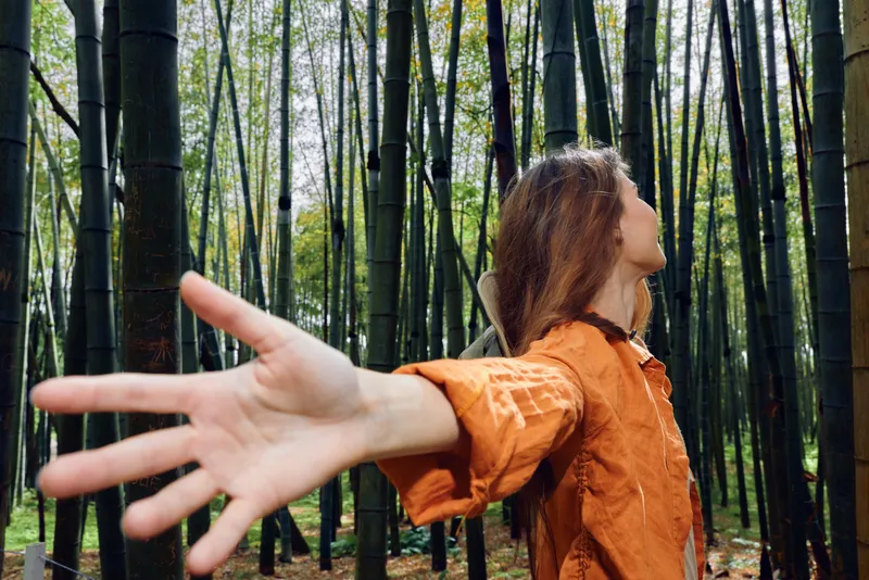 Woman in bamboo forest with outstretched hand among tall trees, portrait in orange shirt, nature scene conveying freedom, exploration and peaceful outdoor moment
