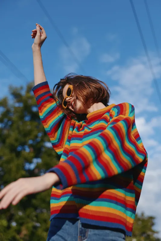 Young woman in her early 20s wearing a rainbow sweater and sunglasses outdoors under blue sky, smiling and dancing with raised arm in casual outfit for lifestyle and fashion.