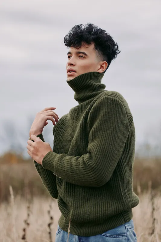 An authentic portrait of a man in an olive green turtleneck sweater stands in an open field, exuding authenticity and credibility through a natural, candid pose and confident gaze.