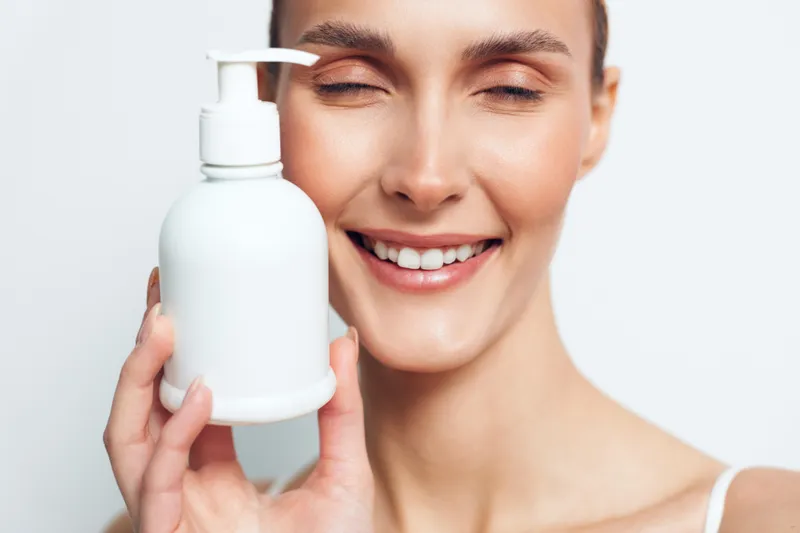 Young woman with clear skin smiles gently while holding a white lotion bottle close to her face against a plain white background.