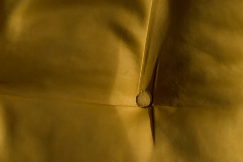 Close-up of a mustard yellow fabric with a single button tuft creating soft folds and shadows on the surface.