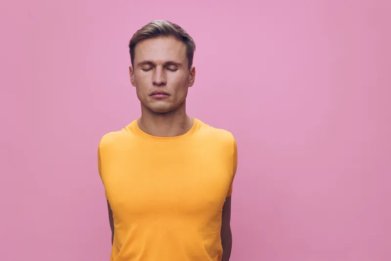 Calm young man meditating closed eyes sleeveless yellow shirt pink background mental health digital detox relaxation concept