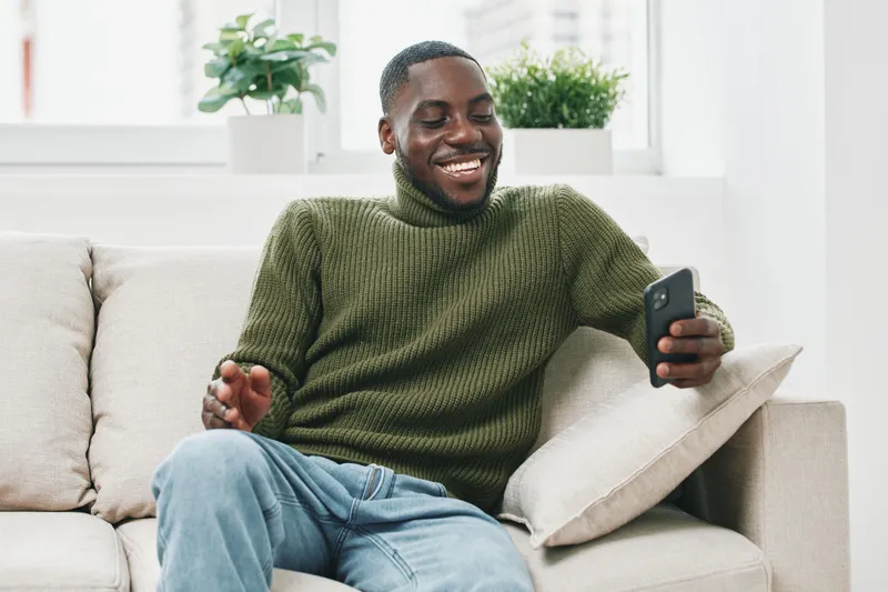 Happy man in a green sweater enjoying his smartphone on a cozy couch in a modern living room, representing joy and relaxation in a bright indoor setting