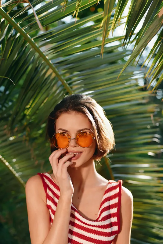 A cheerful young woman wears a striped red and white top and round orange sunglasses as she poses among lush palm leaves, radiating sunny, relaxed style and tropical charm.