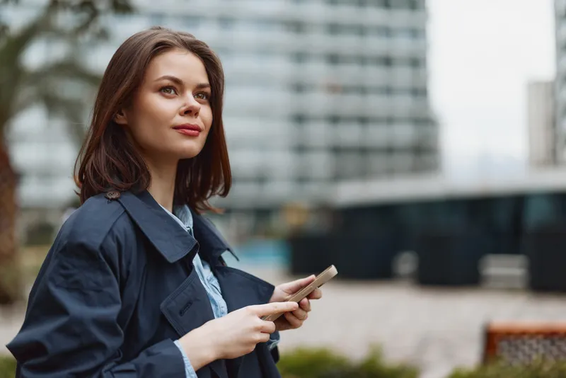 Young woman with brunette hair holds a smartphone outdoors in a city setting while looking thoughtfully into the distance wearing a dark coat and light blouse.