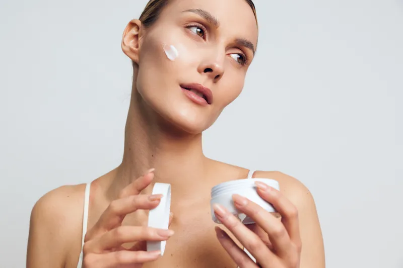 A young woman with clear skin applies white face cream on her cheek while holding a container and a small spatula. The neutral background emphasizes skincare and beauty routine concepts.