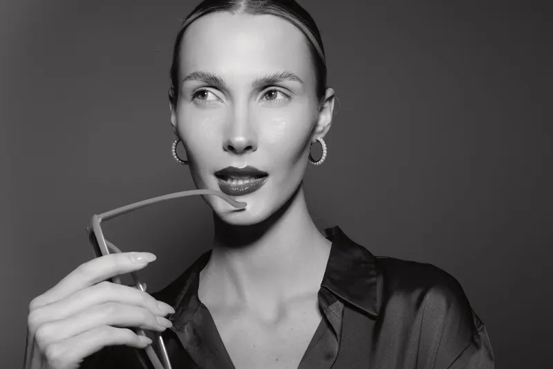 Caucasian woman with fair skin and slicked hair holds eyeglasses near lips, looking away. Studio portrait with dramatic black and white lighting.