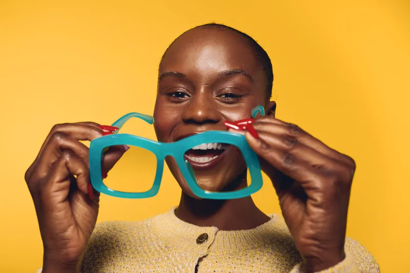 Smiling black woman with dark skin holds oversized blue plastic glasses in front of her face. Yellow background and casual beige sweater create fun and playful mood for lifestyle concept.