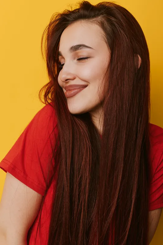 Young woman with long hair and closed eyes smiling gently against yellow background wearing red shirt. Portrait of happy female with natural makeup and relaxed expression.