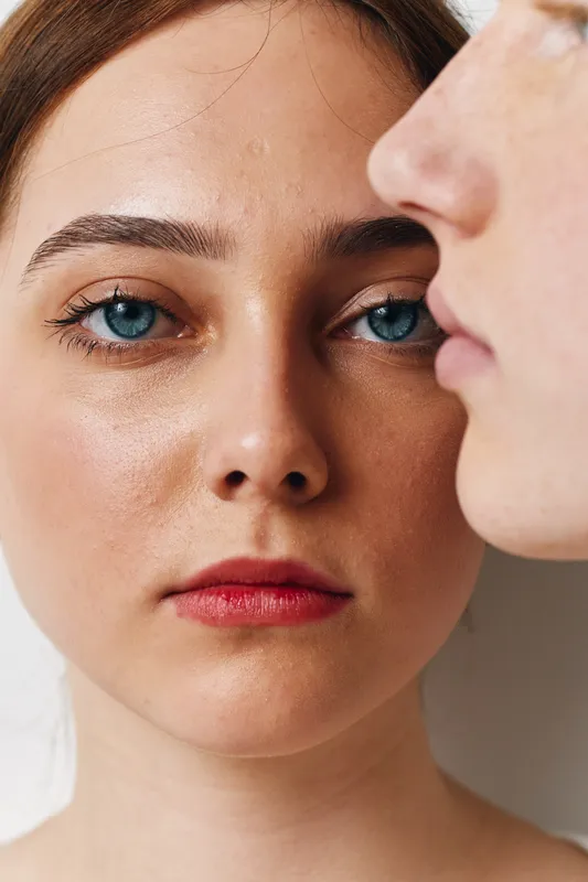 Closeup portrait of woman with blue eyes and red lips looking at camera with man profile blurred in foreground. Natural skin and serious expression in studio light.