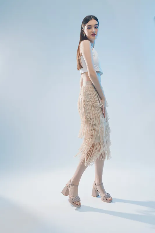 Young woman in a stylish beige fringe skirt and white top poses in high heel sandals against a light background. Concept of woman beauty fashion and elegance.