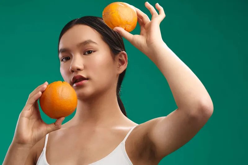 asian woman holding oranges near her temples in a bright studio, a vivid green backdrop emphasizing freshness and vitality, creating a clean, modern mood that highlights fruit and wellness.