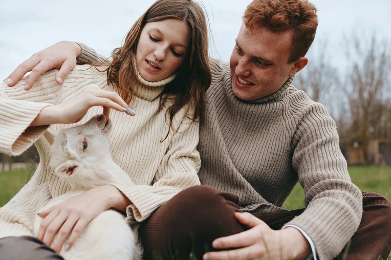 Lifestyle moment outdoors with a couple and a goat kid, tender interaction, warm natural light, soft breeze, cozy sweaters, green grass and calm countryside as background, cheerful mood and relaxed