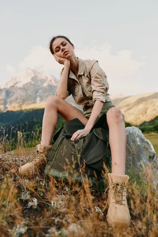 Young woman sits on a rock in a mountain meadow, resting her head on her hand, wearing a khaki shirt, olive shorts, tan boots, and a backpack, soaking in alpine scenery.