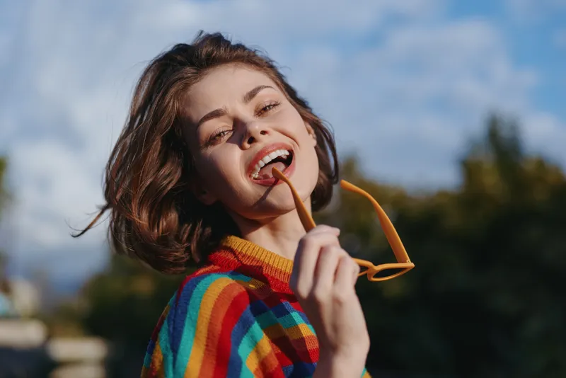 Young woman smiling in a colorful striped sweater, short brown hair, holding sunglasses near her mouth in an outdoor portrait. Casual outfit, joyful lifestyle and sunny mood.
