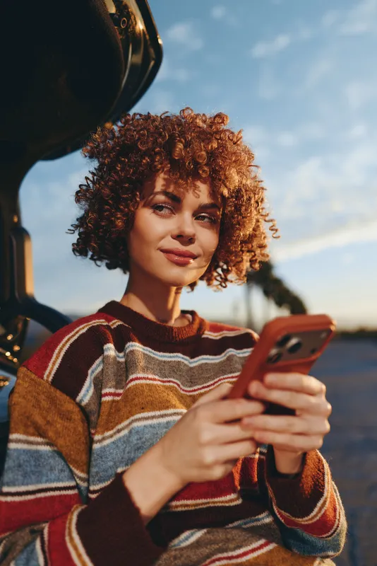 Woman with curly hair outdoors, smiling while holding smartphone near car, wearing striped sweater, warm sunset light, relaxed lifestyle moment, clear sky background soft breeze