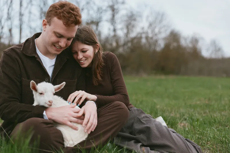 Young couple sitting on grass in casual clothes gently holding a baby goat outdoors in a natural setting with soft light and overcast sky.