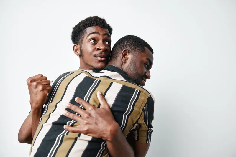 Two black men hugging each other with joy and excitement on white background. African ethnicity, dark skin, friendship and celebration with casual striped shirt.