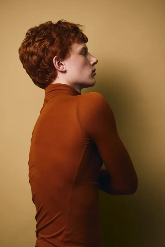 Young man with red hair and pale skin wearing a brown turtleneck sweater, posing sideways against a beige background with folded arms in a studio setting.