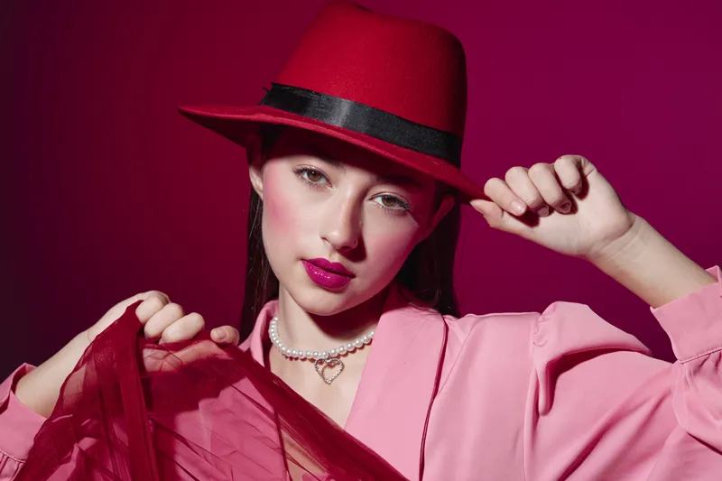Fashion model in a stylish red hat and pink coat poses confidently against a vivid crimson background. Lipstick and pearls highlight her edgy, artistic beauty concept.
