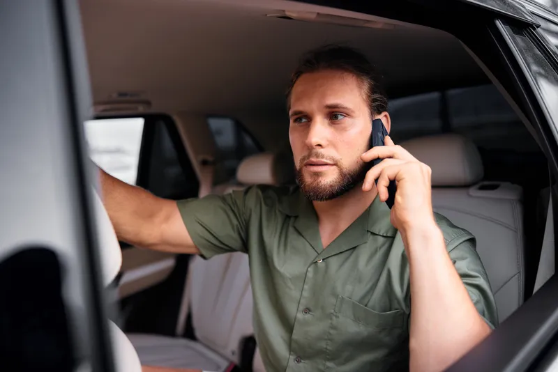 Молодой человек в повседневной зеленой рубашке сидит в автомобиле и разговаривает по телефону