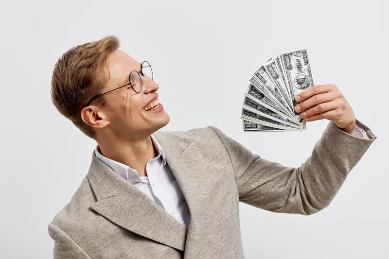 Confident smiling man with glasses holding fan of hundred dollar bills while looking to side dressed in beige blazer and white shirt against plain light background studio portrait. People lifestyle co