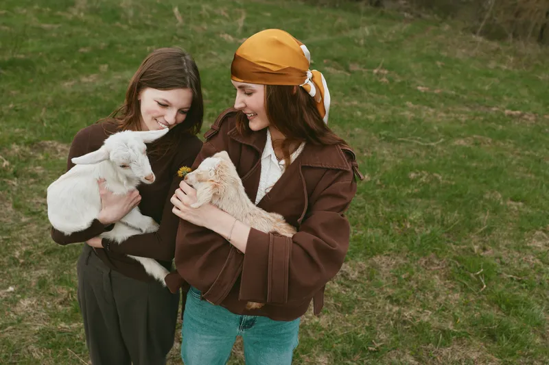Two women outdoors share a gentle moment with young goats on a grassy hillside, showcasing rural lifestyle, animal care, and bonding. Warm natural light highlights curiosity and tenderness.