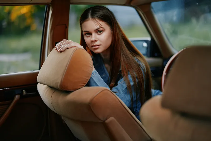 Portrait of a young woman with long hair in a vintage car, showcasing a calm expression and casual denim wear against a soft green background.
