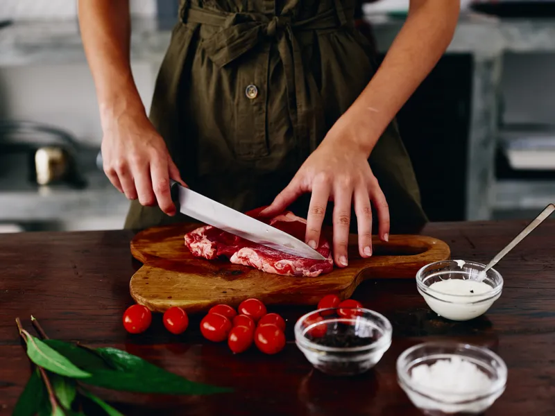 Женщина с ножом в руке нарезает стейк из мяса для жарки на кухне