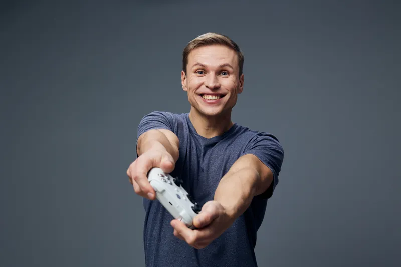 Smiling young man holding game controller extend arms cheerful expression relaxed casual clothing studio portrait gray background People lifestyle concept