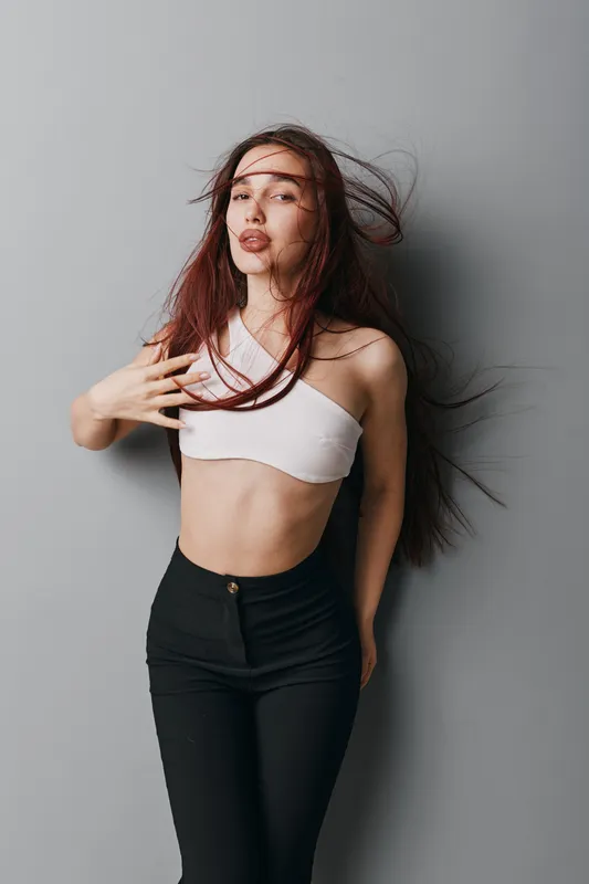 Young woman with long hair posing against gray background wearing white crop top and black pants, fashion portrait with natural makeup and casual style in studio lighting.