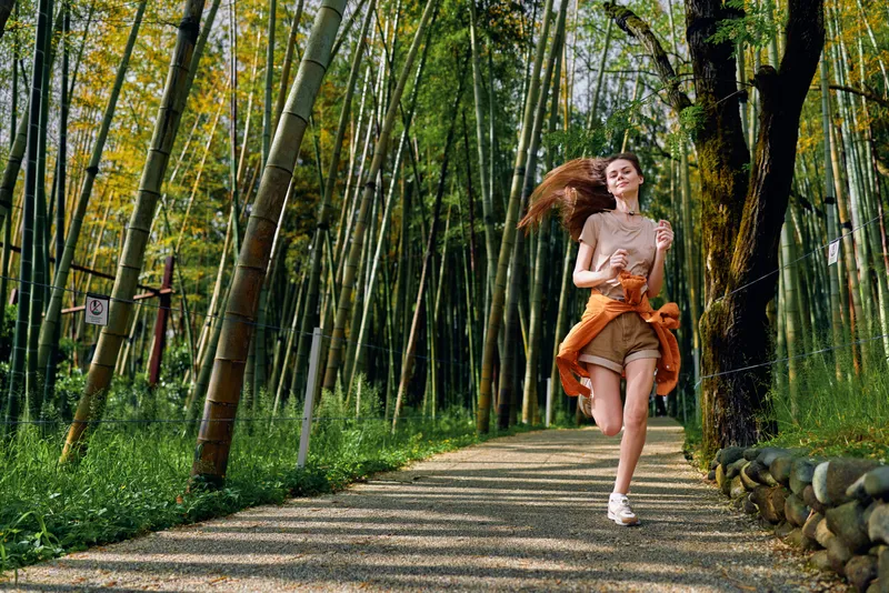 Woman running on a gravel path through a bamboo forest, jogging on a scenic trail in nature for exercise; female runner in casual sportswear enjoys outdoor fitness and sunlit green trees.