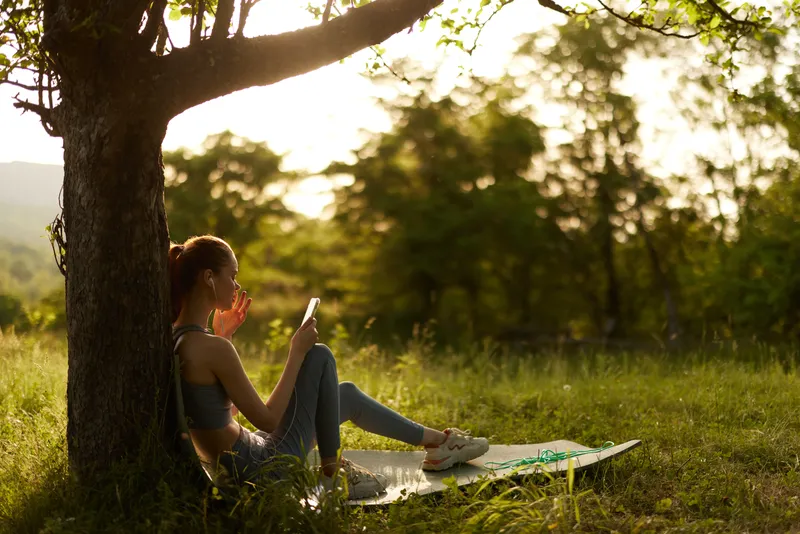 Relaxed young woman reading under a tree in a sunny park, surrounded by greenery, showcasing a calm and peaceful mood during a warm day outdoors