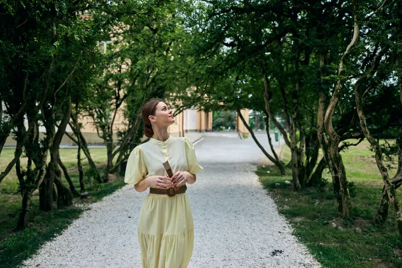 Woman in a yellow dress walks along a gravel path in a lush garden, sunlight filters through trees, creating a serene outdoor scene full of calm and light