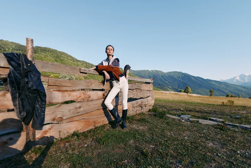 Woman leaning on wooden fence in a field with distant mountains, countryside portrait of outdoors fashion model wearing jacket and boots, relaxed pose in natural landscape.
