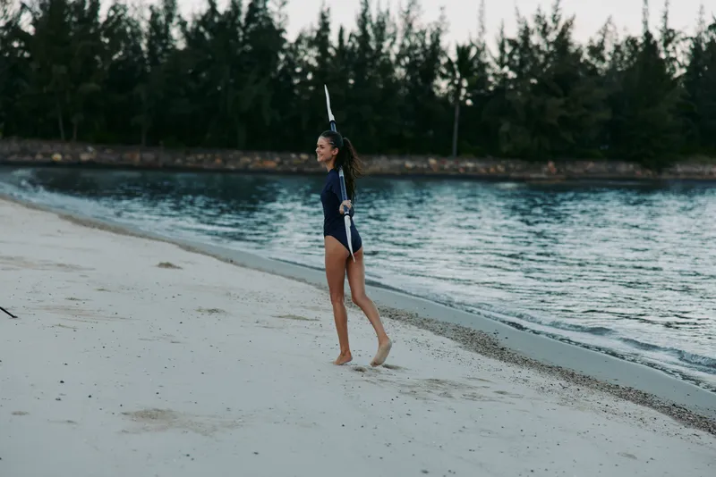 A young girl in a swimsuit walks along a pale sandy beach beside a calm blue sea, wearing a playful unicorn horn headband as gentle waves wash over her feet.