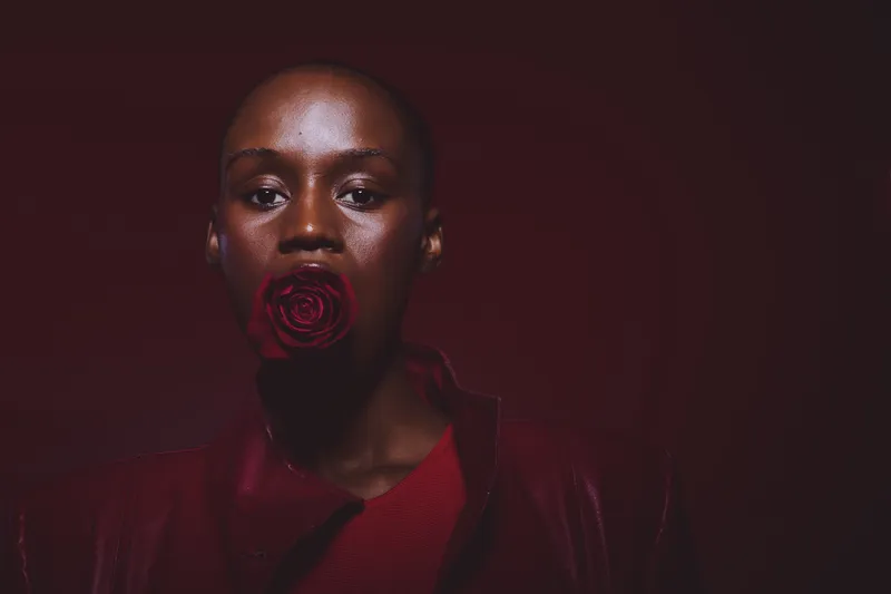 Portrait of a dark skinned African woman with a red rose in her mouth wearing red clothing. The moody lighting and dark background create a dramatic and artistic concept of beauty and expression.