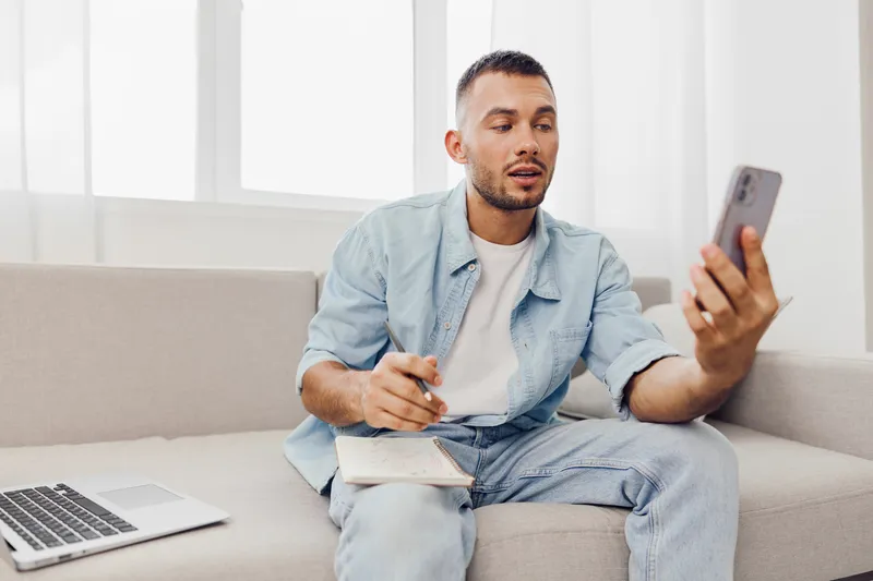 Engaged man using smartphone while sitting on the sofa, dressed casually in a light blue jacket and enjoying his time. Perfect for modern lifestyle themes.