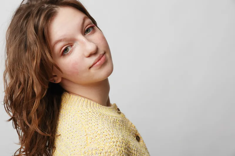 Young woman with curly brown hair wearing yellow knitted sweater looks softly at camera on grey background. Concept of natural beauty, casual style, and gentle expression in studio portrait.