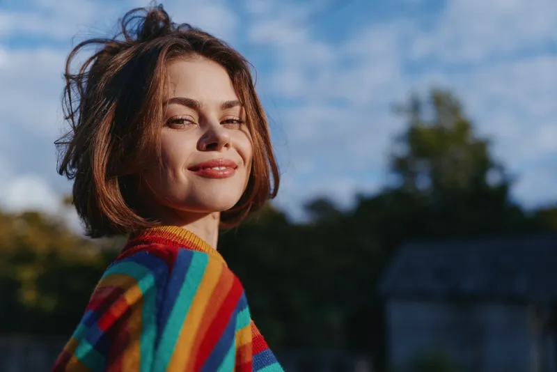 Woman portrait smiling in a colorful rainbow sweater outdoors in warm sunlight, short bob hair and natural skin texture, confident expression with blurred background and blue sky.