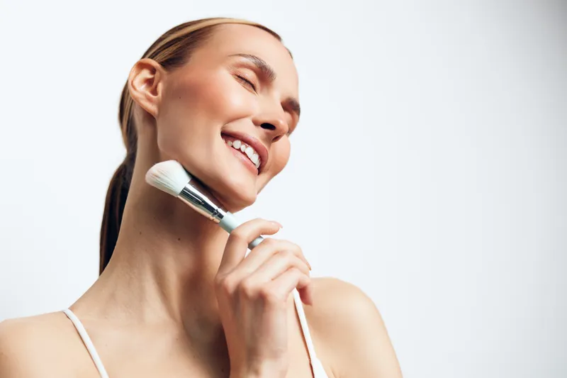 Young woman with clear skin smiling gently while applying makeup powder with a brush, bright studio lighting, minimalistic white background, natural beauty concept.