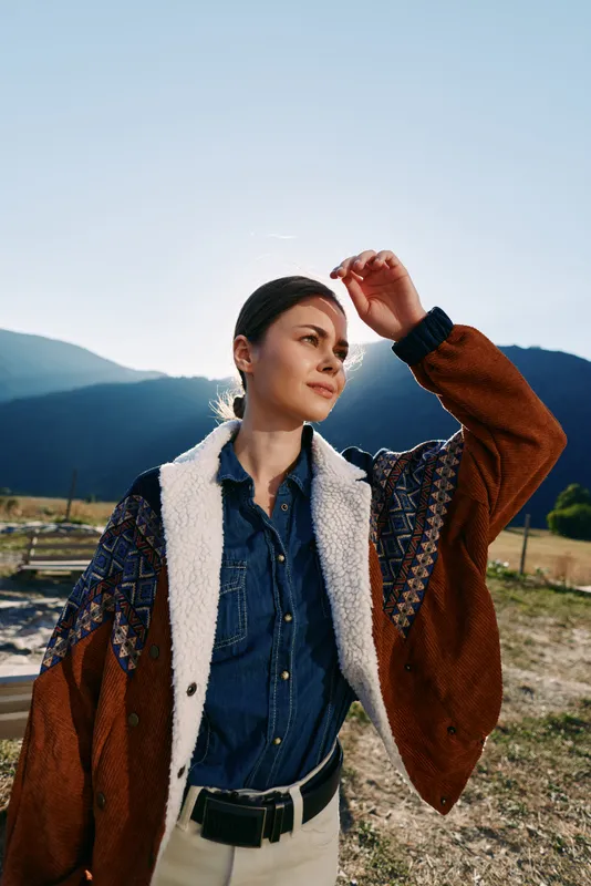 Woman portrait in mountains outdoors with sunlight, wearing denim shirt and cozy cardigan, hand raised to shade eyes. Scenic countryside scene evokes travel, fashion and peaceful adventure.