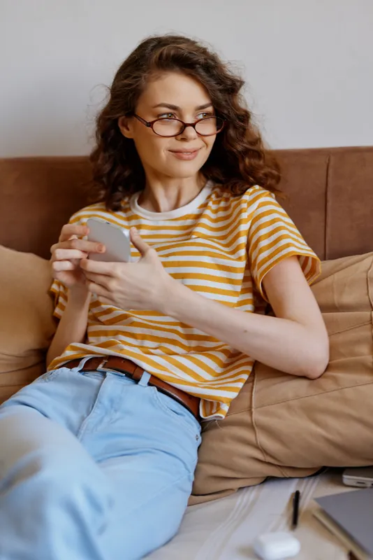 Casual woman at home sits on a couch, wearing striped shirt and glasses, smiling as she uses a smartphone, relaxed mood, daytime indoor setting, cozy living room scene.